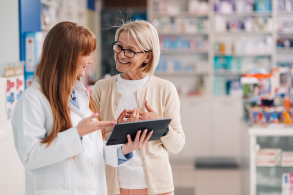 Apothekerin im weißen Kittel erklärt einer Kundin Informationen auf einem Tablet in einer modernen Apotheke.