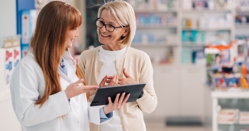 Apothekerin im weißen Kittel erklärt einer Kundin Informationen auf einem Tablet in einer modernen Apotheke.